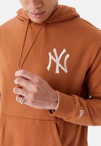 Sudadera marrón con bolsillo frontal, capucha con cordón y logo blanco de Nueva York. Puños y dobladillo acanalados, material suave con una textura lisa.