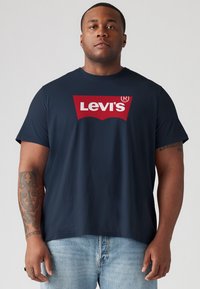 Sötétkék pamut póló, nagy piros Levi's logóval a mellkasán. Rövid ujjú és kényelmes fazonú. Világoskék farmer rövidnadrág viselve.