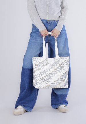 Henkilö, jolla on kaksiväriset leveälahkeiset farkut ja vaalea neuletakki, pitäen valkoista punottua olkalaukkua, jossa on punotut kahvat.