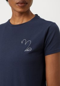 Námornícky bavlnený tričko s okrúhlým výstrihom a krátkymi rukávmi. Obsahuje trblietavý strieborný dizajn z rhinestone na ľavej hrudi.