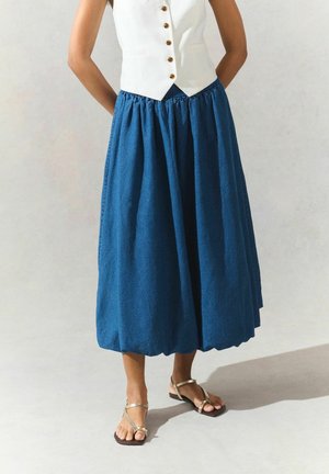 Mujer con chaleco blanco abotonado, falda midi azul voluminosa y sandalias planas doradas con tiras, de pie contra un fondo claro y sencillo.