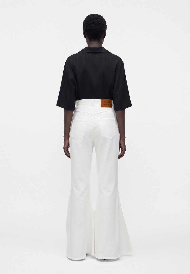 Person with short hair wears black cropped sleeve shirt and high-waist white flared pants with leather patch on back waistband.