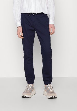 Hombre con pantalones jogger azul marino, zapatillas beige y grises, y una camisa blanca de manga larga con finas rayas azules, de pie sobre un suelo gris.