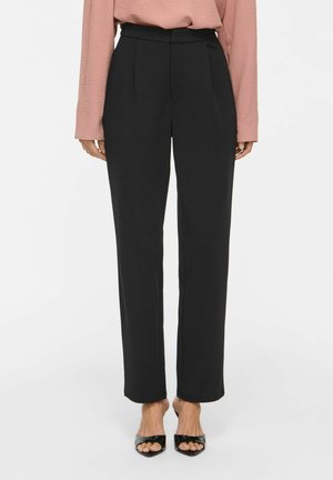 Femme portant un pantalon noir taille haute à jambes droites, associé à un chemisier rose à manches longues et des talons ouverts noirs.
