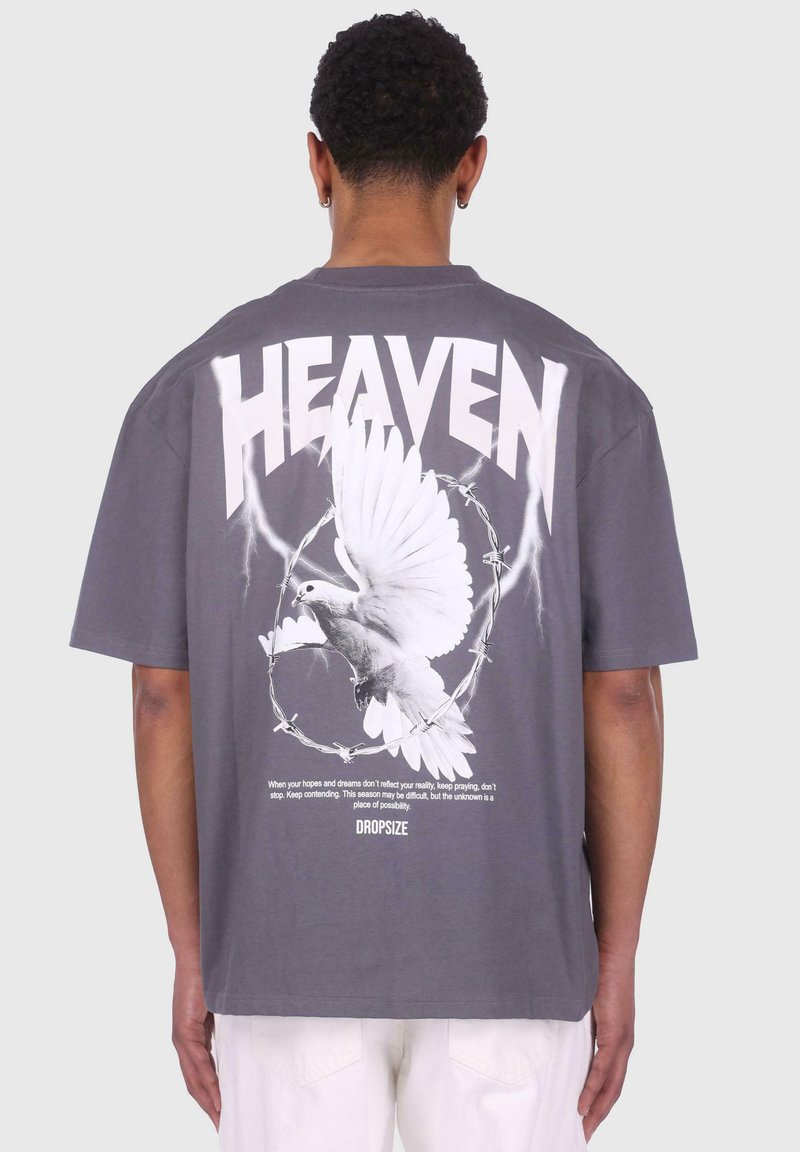 Tricou supradimensionat gri cu grafice albe, inclusiv "HEAVEN", o porumbel și sârmă ghimpată. Textul deasupra și dedesubtul porumbelului conține fraze motivaționale.
