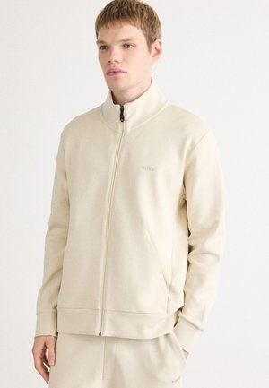 Ung man klädd i beige zip-up jacka och matchande byxor, stående mot en vit bakgrund, med händerna i fickorna.