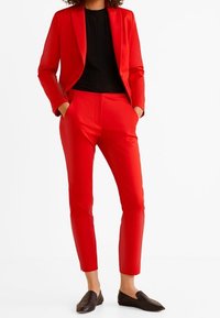 Rood op maat gemaakte blazer met bijpassende smalle broek, gemaakt van gladde stof. Draagt over een zwart topje, met zwarte puntige ballerina's.