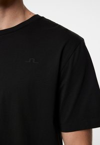 Mustat pyöreän kaula-aukon t-paita, joka on valmistettu sileästä kankaasta. Etupuolella on huomaamaton musta logo vasemmalla ylhäällä. Lyhyet hihat, joissa on siistit saumat.