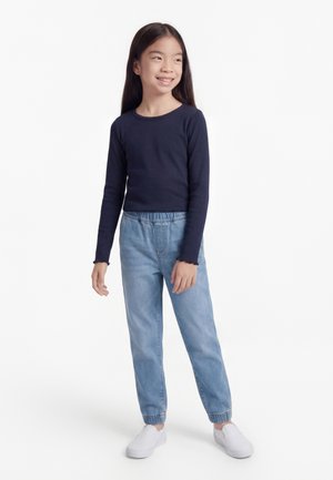 Noor tumehalli pikakarvaga tüdruk, kes kannab tumesinist pika varrukaga särki, helesiniseid elastse vöökohaga teksapükse ja valgeid libisevaid jalanõusid, seisab ja naeratab.