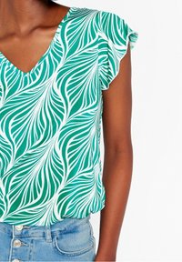 Zielono-biała bluzka z dekoltem w kształcie litery V, zdobiona wzorem liści i falbankowymi rękawami. Noszona z jasnoniebieskimi dżinsami z wysokim stanem.