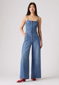 Denim kombinezons ar pieguļošu augšu, platu kājām, regulējamām šaurām uzsvaru siksnām un priekšējo rāvējslēdzēju. Gaismas zilā krāsā, neformāla tekstūra.