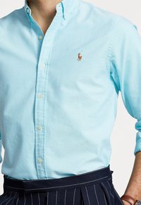 Ljusblå skjorta med knäppning i bomull, med klassisk krage och en liten broderad logotyp på bröstet. Strukturerad tyg.