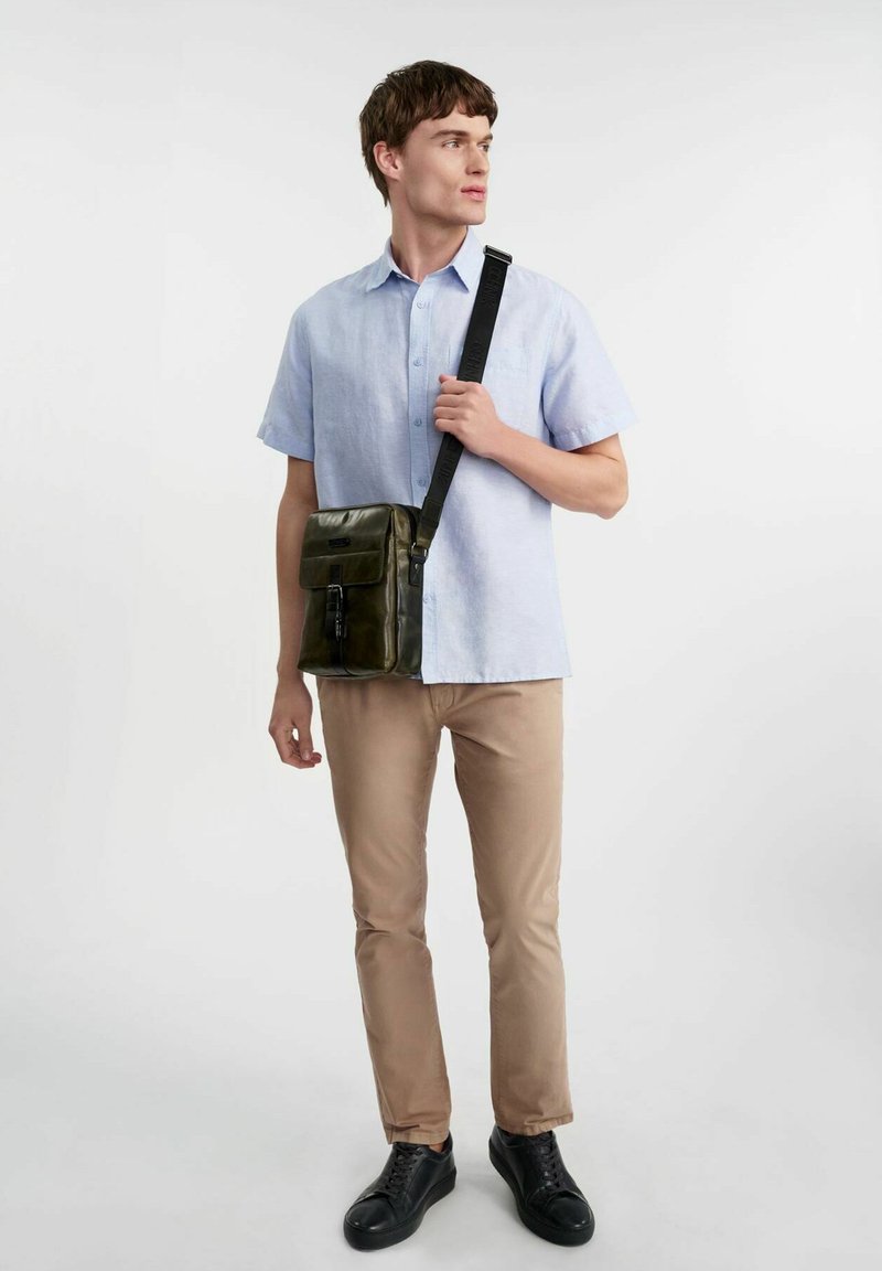 Zielona skórzana torba na ramię z klapą, czarnym regulowanym paskiem i przednią kieszenią na zamek. Mężczyzna ma na sobie jasnoniebieską koszulę z krótkim rękawem oraz beżowe spodnie.