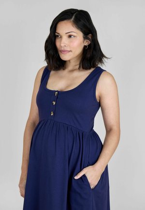 Kvinde med skulderlangt mørkt hår iført en ærmeløs marineblå kjole med knapper, stående med den ene hånd i lommen og kiggende til siden.