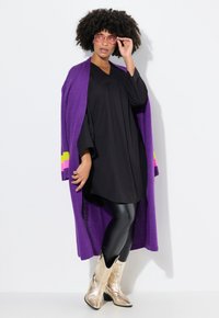 Sort tunika top, lilla cardigan med pink og grønne accenter, sorte leggings og metaliske guld cowboy støvler. Glatte teksturer og livlige farver.