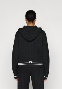 Hanorac tricotat negru cu un model de cablu și glugă. Are un accent cu dungi albe la talie. Designul este de tip relaxed fit.