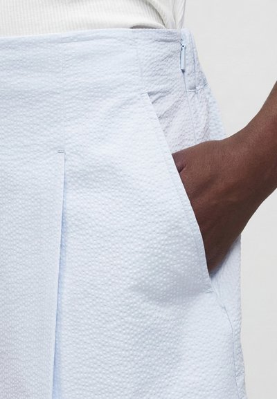 Χέρι με σκούρο χρώμα δέρματος στην τσέπη ανοιχτό μπλε υφασμάτινης φούστας, με λευκό ριμπ πλεκτό τοπ ορατό στη μέση, πάνω σε απλό φόντο.