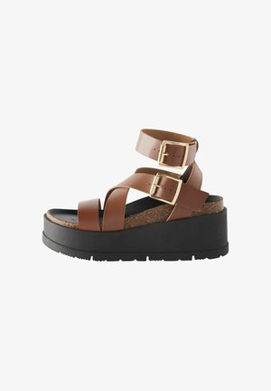 Bruine faux leren platform sandalen met twee enkelbandjes, gouden gespen en een gestructureerde kurken voetbed, geplaatst op een zwarte rubberen zool.