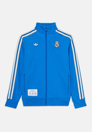 Veste zippée bleue avec des rayures blanches et dorées, un col haut et les logos d'Adidas et du Real Madrid. Fabriquée dans un matériau lisse.