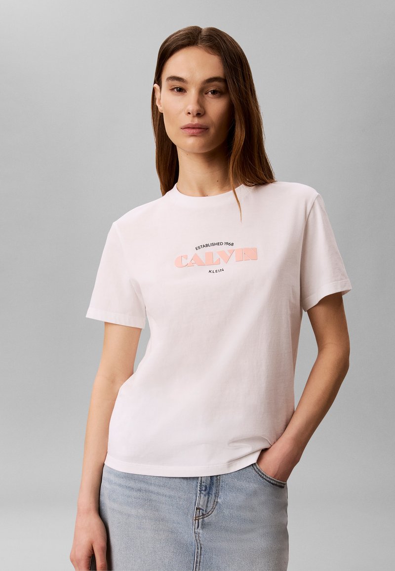 Femme portant un t-shirt blanc Calvin Klein avec un logo rose, associé à une jupe en jean bleu clair, debout devant un fond gris uni.