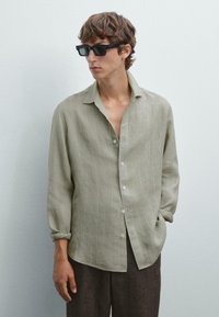 Camicia a maniche lunghe verde chiaro con strisce verticali bianche, tessuto leggero, con bottoni davanti, vestibilità comoda, abbinata a pantaloni scuri e occhiali da sole neri.