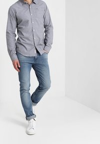 Šedá košile na knoflíky s kapsou, kombinovaná s světle modrými džínami a bílými teniskami. Košile má límec.