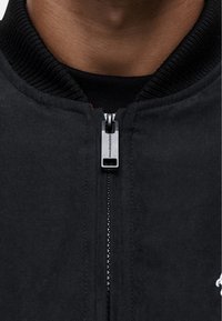 Giubbotto bomber nero con colletto a coste, dotato di una zip argentata e di un logo ricamato bianco sul lato sinistro vicino al collo.