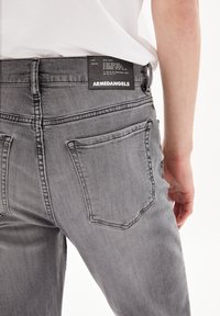 Šedé džíny z denimu s obepínajícím designem. Mezi prvky patří ploché zadní kapsy a černá značka. Textura je hladká s klasickým finišem.