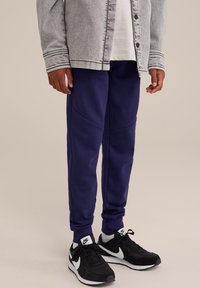 Mornarsko modri trenirke s ožjim krojem, ki so opremljene z teksturiranimi paneli, skupaj z črnimi športnimi čevlji Nike z belimi detajli.