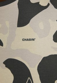 Beige ir juodos spalvos kamufliažo raštuotas audinys su lygiu paviršiumi. Yra prekės ženklo „CHASIN'“ pavadinimas išryškintas storu juodu šriftu.