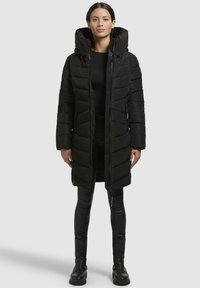 Black puffer coat with a hood, featuring horizontal stitching, front zipper, and side pockets. Worn over a black top and fitted pants.
