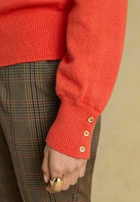 Korallfarbener gestrickter Pullover mit gerippten Bündchen und drei goldenen Knöpfen am Ärmel, kombiniert mit karierter Hose im Braun- und Grünton.