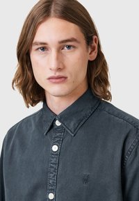 Camicia button-up grigia scura con colletto a punte, bottoni bianchi e un piccolo logo ricamato sul lato sinistro del petto. Tessuto morbido.