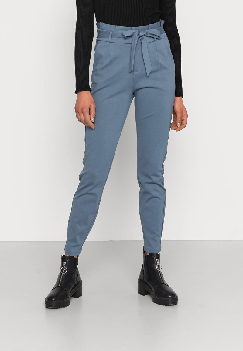 Femme portant un pantalon paperbag bleu taille haute avec une ceinture nouée, un top noir à manches longues en maille côtelée et des bottines noires zippées à l'avant.