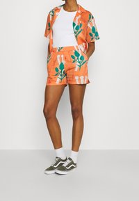 Kortärmad skjorta och shorts-set i orange med gröna växtmönster. Bomullsmaterial, avslappnad passform, kombinerat med vita strumpor och gröna sneakers.