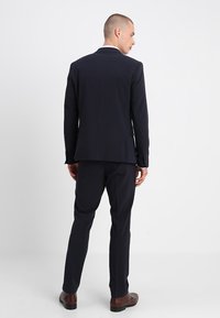 Marineblaues Anzug, taillierte Passform, einreihige Jacke, Reverskragen, flache Hose, glatter Stoff, Knopf-Akzente, kombiniert mit braunen Schuhen.