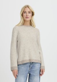 Beżowy sweter w paski z długimi rękawami, okrągłym dekoltem i strukturalnym wykończeniem. Subtelna zmiana koloru na dolnej krawędzi i mankietach. Noszony z jasnymi dżinsami.