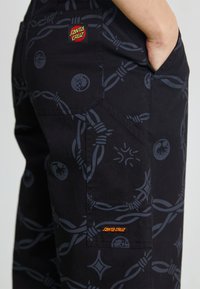 Pantaloni cargo neri con modelli grafici grigi sottili, compresi design annodati e accenti circolari, con un logo "Santa Cruz" rosso e arancione.
