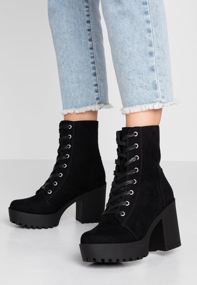 Bottes à plateforme en daim noir avec talons épais et lacets à l'avant, portées avec un jean denim bleu clair coupé au-dessus de la cheville.