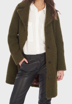 Kvinde iført en åben olivengrøn tekstureret frakke, hvid skjorte med knapper og sorte læderbukser, med den ene hånd i lommen.