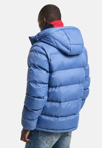 Modrá péřovka s kapucí, prošívaný design a syntetický materiál. Má hladkou texturu a stáhnutelný lem pro teplo.