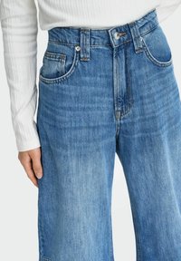 Plave traperice visokog struka, izrađene od denima, s klasičnim dizajnom od pet džepova, kopčanjem na dugme i ravnim krojem.