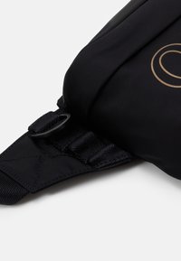 Sangle en tissu noir avec renforcement cousu et anneau en D en plastique, fixée à un sac noir, avec une partie d’un logo circulaire doré visible.