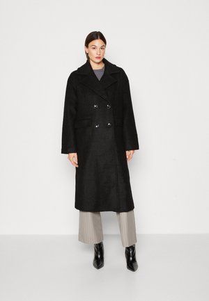 Svart ullkappa med dubbelknäppt design, stora slag och framträdande knappar. Har sidofickor och en medellång längd.