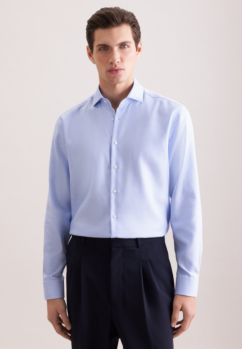 Homme portant une chemise habillée à manches longues à carreaux bleu clair, rentrée dans un pantalon noir taille haute plissé, debout devant un fond uni.