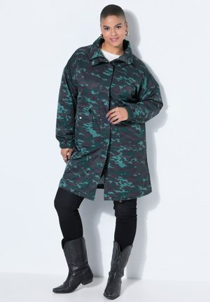 Žena nosící dlouhý kabát s maskáčovým vzorem v tmavě zelené a černé barvě, černé legíny a černé kovbojské boty, stojící na bílém pozadí.