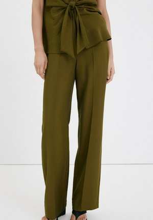 Mujer vistiendo pantalones de pierna ancha color verde oliva combinados con una blusa a juego que presenta un detalle de nudo frontal, posando sobre un fondo blanco liso.