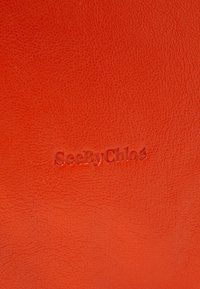 Materiale in pelle arancione con texture che presenta un logo impresso "SeeByChloé" per un dettaglio di branding. Superficie liscia con una leggerissima venatura.