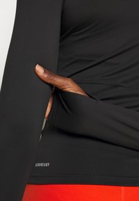 Dłoń w otworze na kciuk czarnej długiej koszulki sportowej oznakowanej "AEROREADY", noszonej z jasnoczerwonymi dołem.