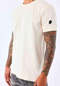 Camiseta de manga corta beige con cuello redondo, tejido texturizado y un parche de logo negro en la manga. Diseño ajustado.